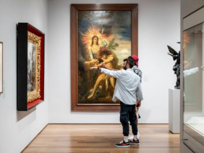 In the corner of a gallery, two people stand near a painting; one of them points toward it.