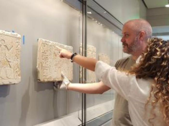 A woman points to a relief fragment, which is being held up by a man next to her, in an open wall case.