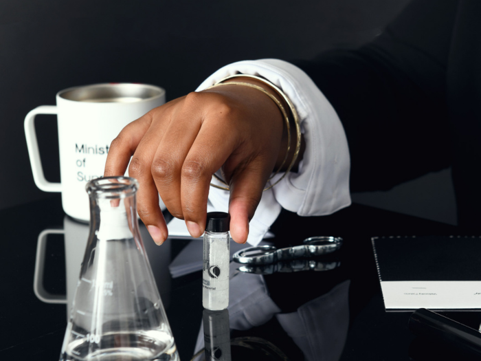 A dark brown hand in formal attire is holding a 5 gram vial, with a black lid and the Mbadika logo on the container, above a black laboratory table. The background showcases a modern laboratory with a flask, fabric sample, and a white metal cup.