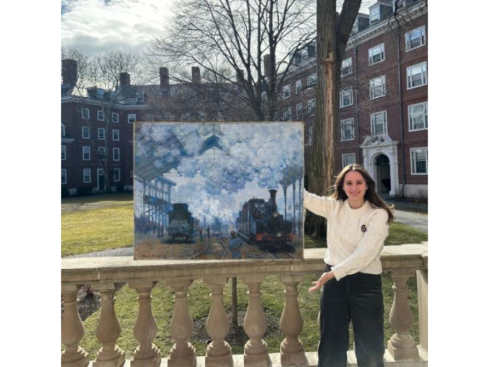 A smiling young woman stands in a campus quad gesturing toward a painting that appears to be floating on top of the balustrade behind her. The brushstrokes coalesce into a bluish scene of trains at a smoke-filled station.