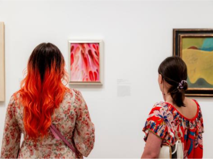 Two women view framed artworks in a gallery setting.