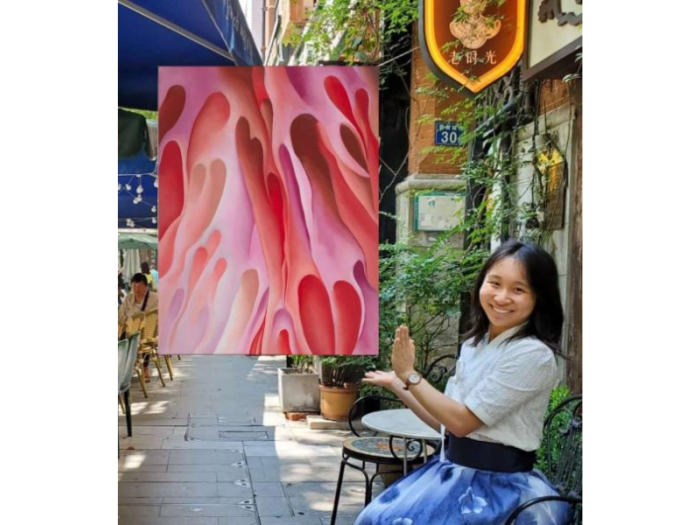A smiling young woman seated in a cafe terrace gestures toward a nearly abstract painting that appears to float beside her. It represents a close-up view of folding red and pink forms.