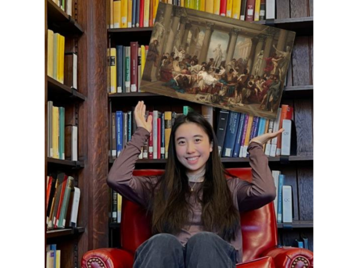 A smiling young woman, wearing a brown long-sleeve shirt and dark pants, sits in a red armchair in a library, gesturing toward a painting that appears to float overhead. In the painting, people in Roman clothes gather in a lofty and statue-filled classical setting.