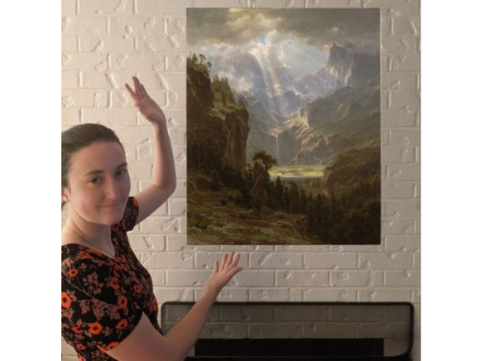 A young woman in a black patterned dress stands by a mantle gesturing toward a painting that appears to be floating. In the painting, beams of heavenly light illuminate a lush valley nestled in a mountain range.