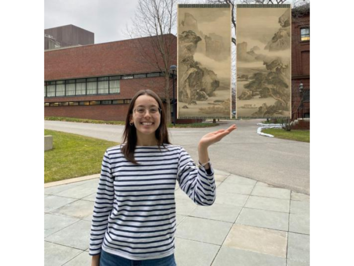 A smiling young woman, standing near campus buildings, gestures toward a pair of hanging scrolls that appear to float. They represent a dramatic, misty landscape. She wears a striped long-sleeve T-shirt and blue jeans.