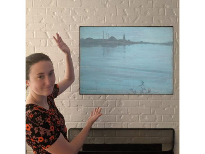 A young woman gestures toward a floating landscape painting over a mantle. The work is an almost abstract composition in silvery blues and grays.