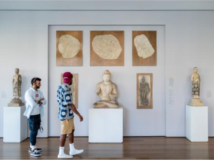 In a gallery space, two people walk toward a sculpture of a large Buddha in a seated position on a pedestal. Surrounding the Buddha are framed works on the wall and two pedestals, each with standing sculptural figures.