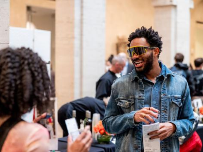 In a courtyard, a man wearing sunglasses and a denim shirt and jacket is smiling and chatting with a person whose back is to the camera. They stand near a long table with refreshments, and a crowd is behind them.