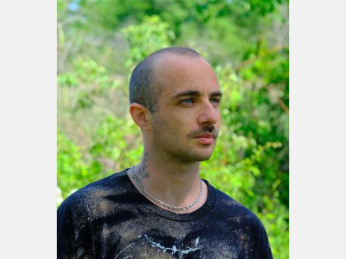 Image features a blue-eyed white-presenting adult male with a moustache and a shaved head looking  off in the distance. In the background are leaves and branches.