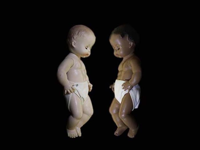 two plastic baby dolls, one with white skin and one with black skin, facing each other against a black backdrop