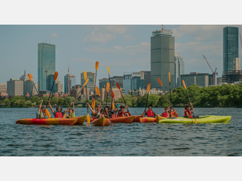 Photo of a large group of youth kayaking on the Charles River with the Boston cityscape behind them.